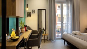 Moderni hotellihuone, jossa on sänky, ruokapöytä tuoleineen, pieni sivupöytä, jossa on pullo ja laseja, ruukkukasvi ja näkymä ulkoiselle parvekkeelle.