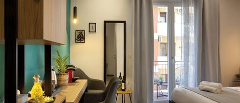 A modern hotel room with a bed, a dining table with chairs, a small side table with a bottle and glasses, a potted plant, and a view of the outside balcony.
