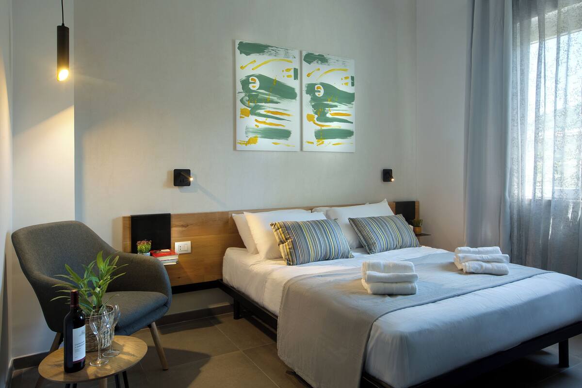 Une chambre d’hôtel moderne, équipée d’un lit, d’une chaise, d’une petite table avec une plante et une bouteille, et ornée d’œuvres d’art au mur.