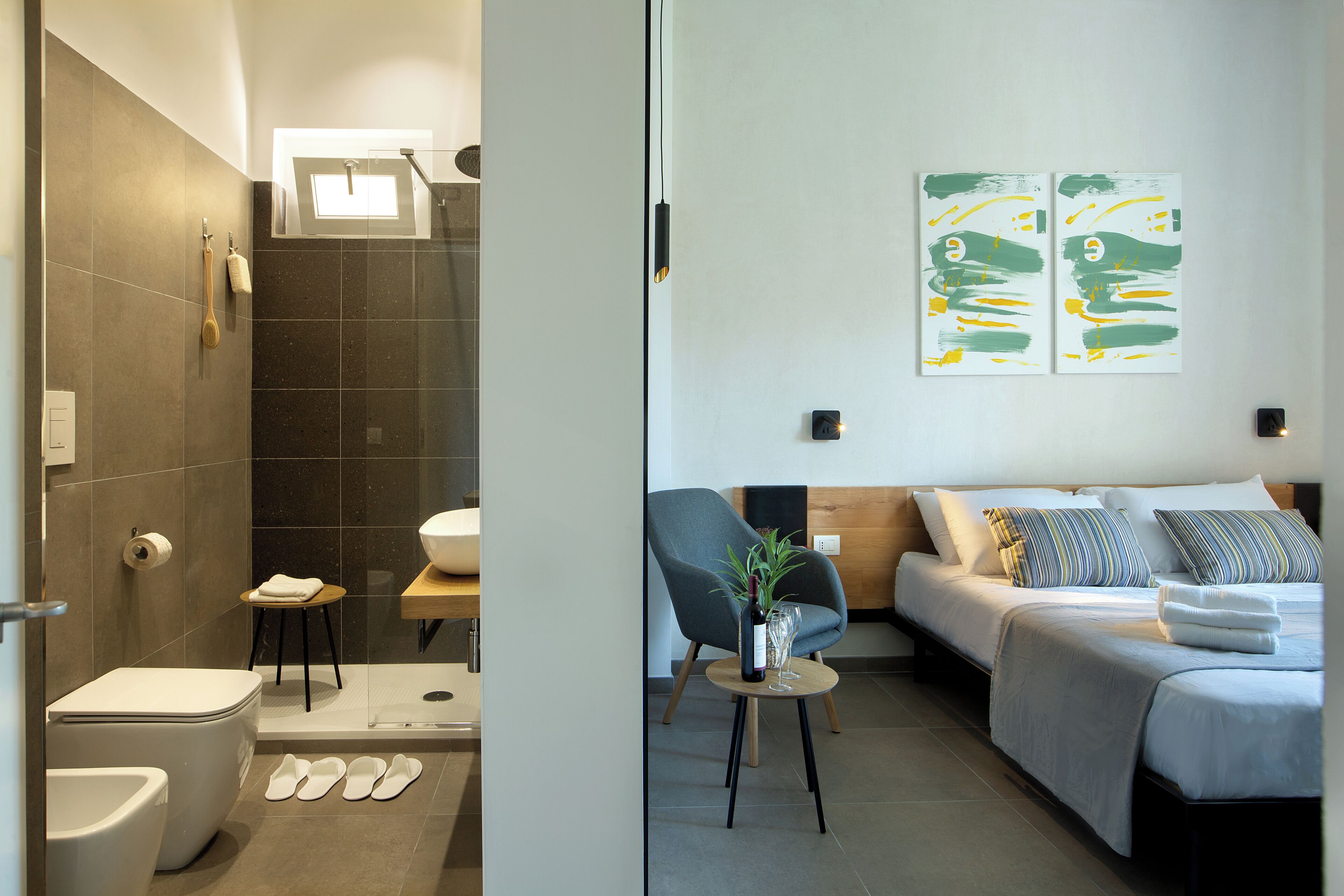 A modern bathroom with a toilet, bidet, and shower. The room features a small table with a plant and a bottle. Adjacent is a bedroom with a bed, pillows, and a bedside table.