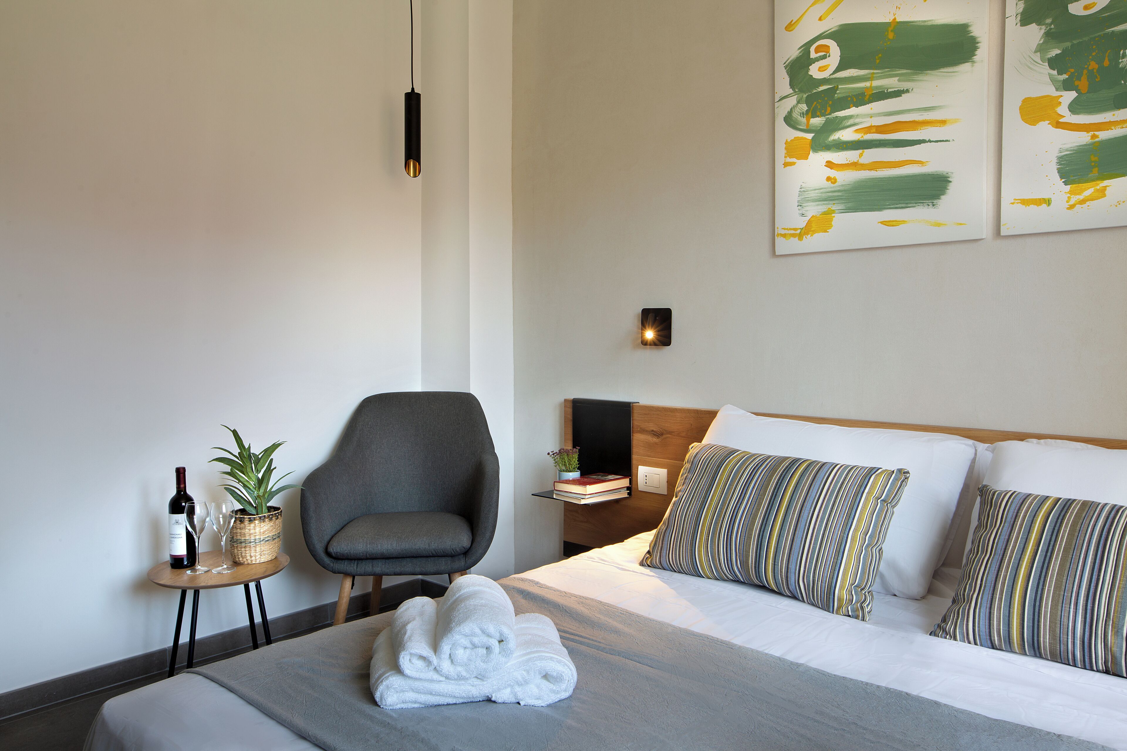 Una camera d'hotel moderna con un letto rifatto, una poltrona grigia, un comodino con una bottiglia di vino e bicchieri, una pianta in vaso e un quadro astratto appeso al muro.