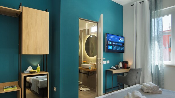 Hotellihuone, jossa on sänky, työpöytä, peili, televisio ja kylpyhuone, joka näkyy avoimesta ovesta.