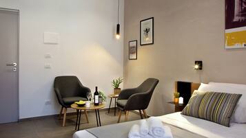 Quarto de hotel com uma cama, duas poltronas, uma mesinha com uma garrafa de vinho, uma planta e um candeeiro fixo na parede.