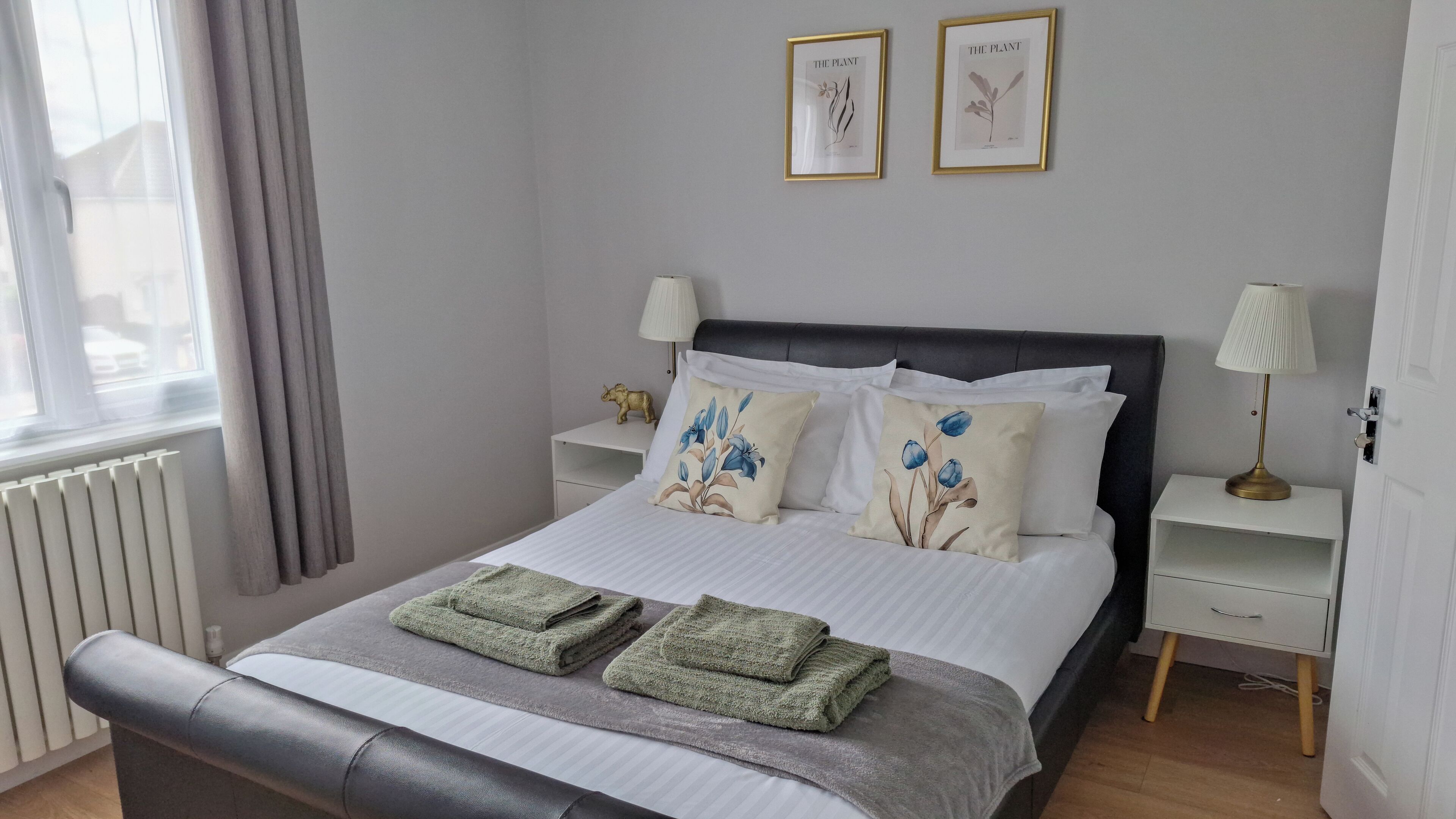 Une chambre à coucher bien rangée, avec un lit, deux tables de chevet, une fenêtre avec des rideaux et deux reproductions botaniques encadrées accrochées au mur.