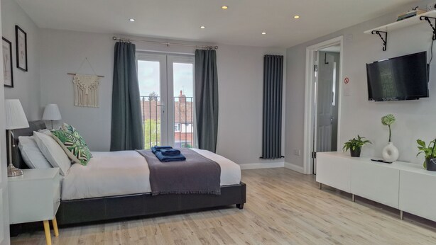 A modern bedroom with a large bed, a TV mounted on the wall, and a balcony with a view of buildings. - NEW 2 bedrooms with private en-suite near Heathrow (Ashford)
