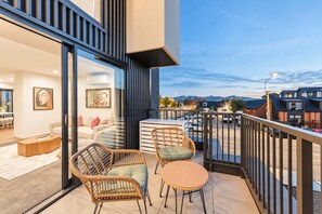 Un balcone con mobili in vimini, una porta vetrata che dà su un soggiorno e vista su un'area di parcheggio e edifici.