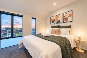 A hotel room with a large bed, a bedside table, a nightstand, and a view of the city through large windows. - Fitzgerald | 3BR + Parking | Sleeps 7 (Christchurch)