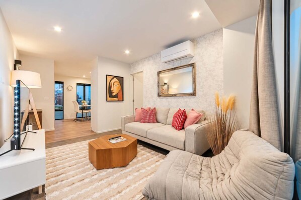 A modern living room with a sofa, a coffee table, and a lamp. There is a painting on the wall and an air conditioning unit above the sofa. - Fitzgerald | 3BR + Parking | Sleeps 7 (Christchurch)