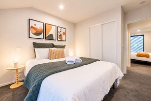 A hotel room with a bed, a bedside table with a lamp, a wardrobe, and a view of the outside. - Fitzgerald | 3BR + Parking | Sleeps 7 (Christchurch)