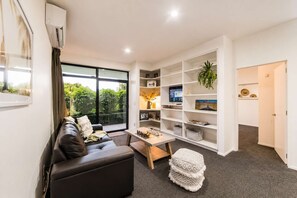 A modern living room with a sofa, coffee table, and a TV. There is a bookshelf with various items and a plant. - City Views | 4BR | Sleeps 10 | Christchurch (Christchurch)