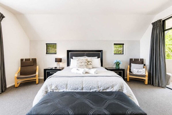 A hotel room with a large bed, two bedside tables, a chair, a window with curtains, and a view of greenery outside. - City Views | 4BR | Sleeps 10 | Christchurch (Christchurch)