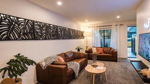 A living room with two brown leather sofas, a round wooden coffee table, a large abstract painting, and a decorative wall panel. - The City Sanctuary | 4BR | 4 Baths | CBD (Christchurch)