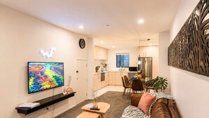 A modern living room with a flat-screen TV, a brown leather sofa, a wooden coffee table, and a dining area with a kitchen in the background.