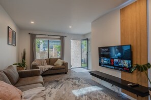 Un salon moderne doté d’un grand téléviseur à écran plat fixé au mur, d’un canapé, d’une table basse et d’une grande fenêtre avec des rideaux.