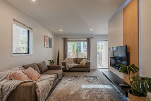 Una sala de estar moderna con un sofá, una mesa de centro y un televisor. Hay un ventanal y una puerta que da al exterior.