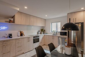 Una cocina moderna con armarios de madera clara, una mesa de comedor de vidrio y una lámpara colgante negra.