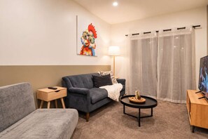 A living room with a grey sofa, a small wooden side table, a coffee table with decorative items, a lamp, and a television on a wooden stand.