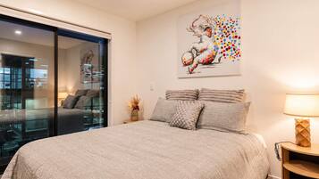 A bedroom with a large bed, a glass door leading to a balcony, a painting on the wall, and a bedside table with a lamp.