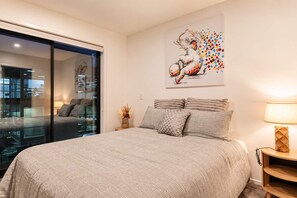 A bedroom with a large bed, a glass door leading to a balcony, a painting on the wall, and a bedside table with a lamp.