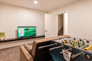 Ein Zimmer mit einem Kicker, einem Fernseher, der einen Sportkanal zeigt, und einer Treppe, die in einen anderen Bereich führt.