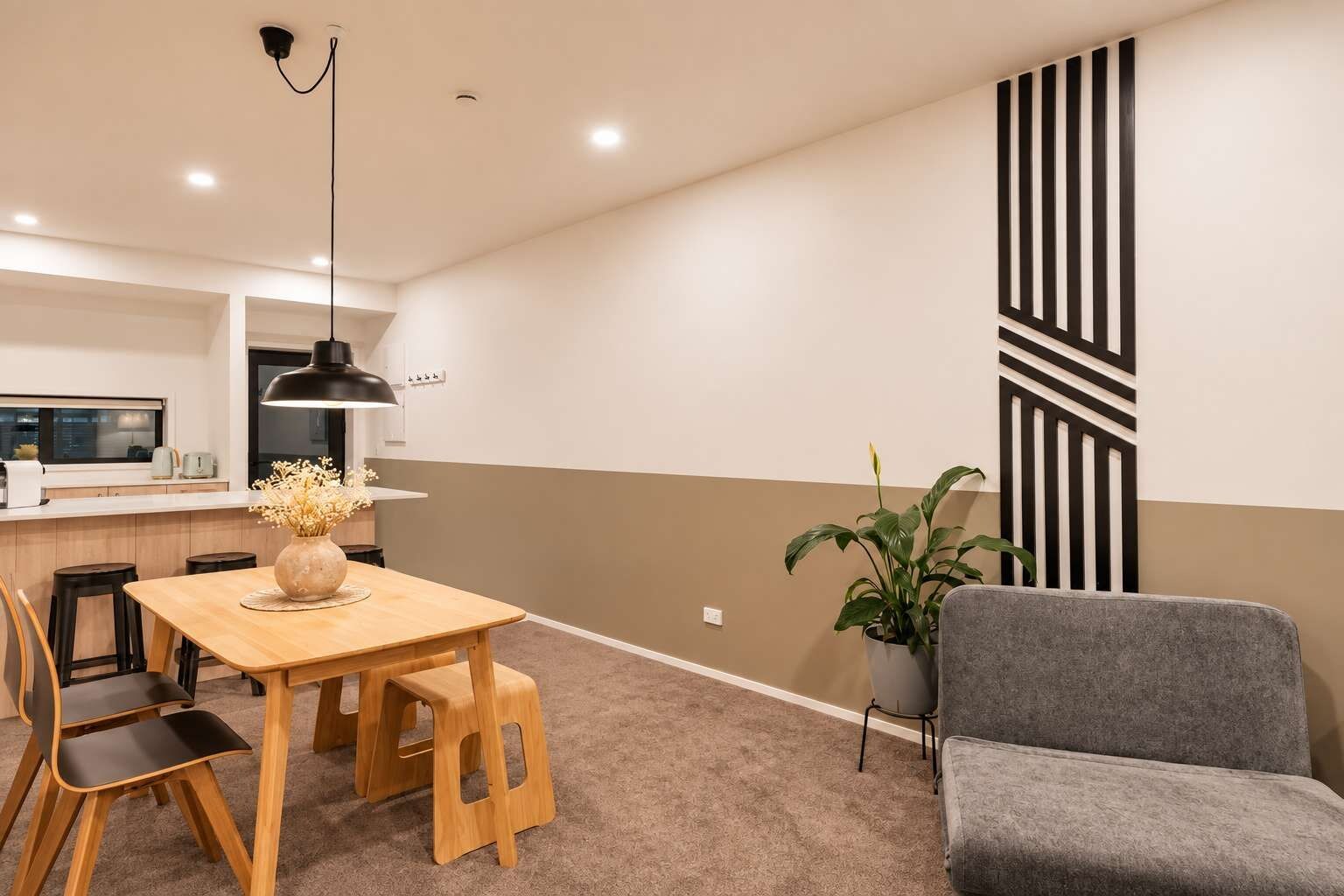 A modern living space with a dining area, a grey armchair, a potted plant, and a kitchen in the background.