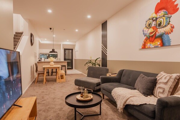 Un salon moderne avec un canapé, une table basse et un téléviseur. On aperçoit à l’arrière-plan un coin repas avec une table et des chaises.