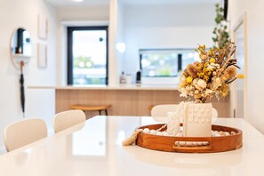 A modern dining area with a white table, a wooden sideboard, and a vase with dried flowers. - Riverside City Base | 4BR + BBQ | Group Stay (Christchurch Central City)