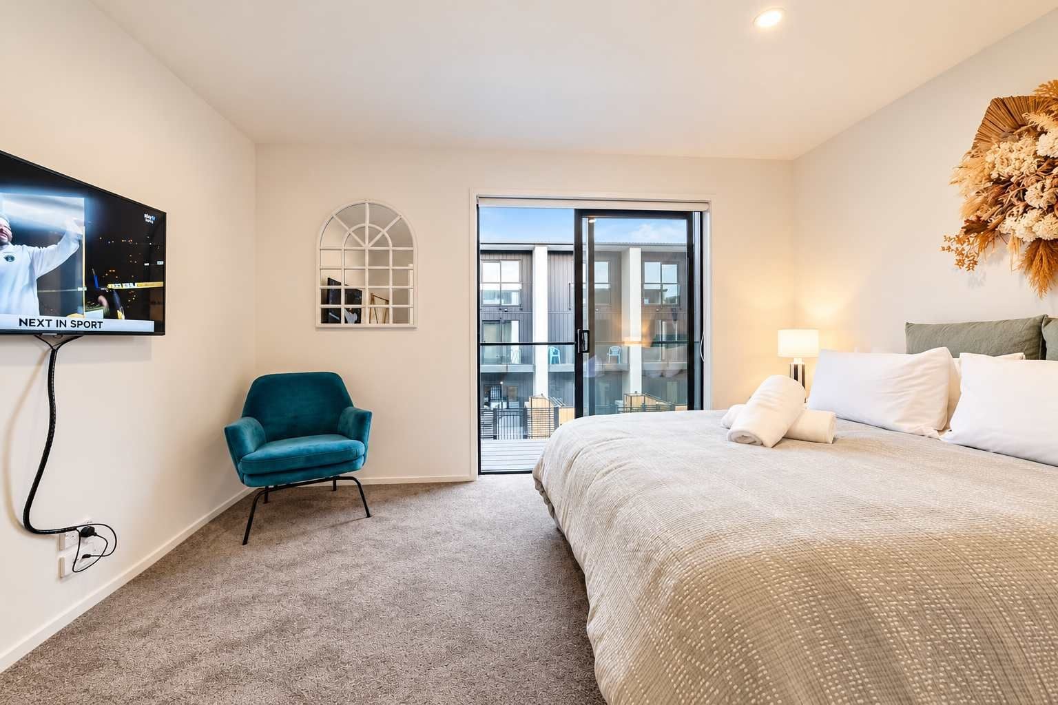 A hotel room with a bed, a teal armchair, a television, and a view through a glass door.