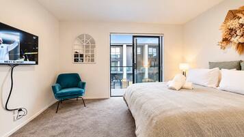A hotel room with a bed, a teal armchair, a television, and a view through a glass door.