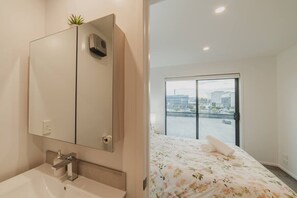 Ein modernes Badezimmer mit großem Spiegel, Waschbecken und Toilette. Ein Bett mit blumigem Überwurf und Blick auf eine Stadtlandschaft durch ein großes Fenster.