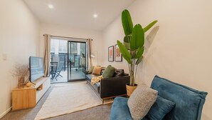 En moderne stue med sofa, fjernsyn, en stor plante og en skydedør til et udendørs område.