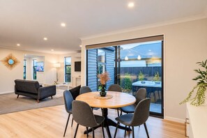 A modern living room with a round wooden table, grey sofas, and a large sliding glass door leading to an outdoor hot tub area. - Wigram 3BR Home | Hot Tub + Games | Sleeps 8 (Christchurch)