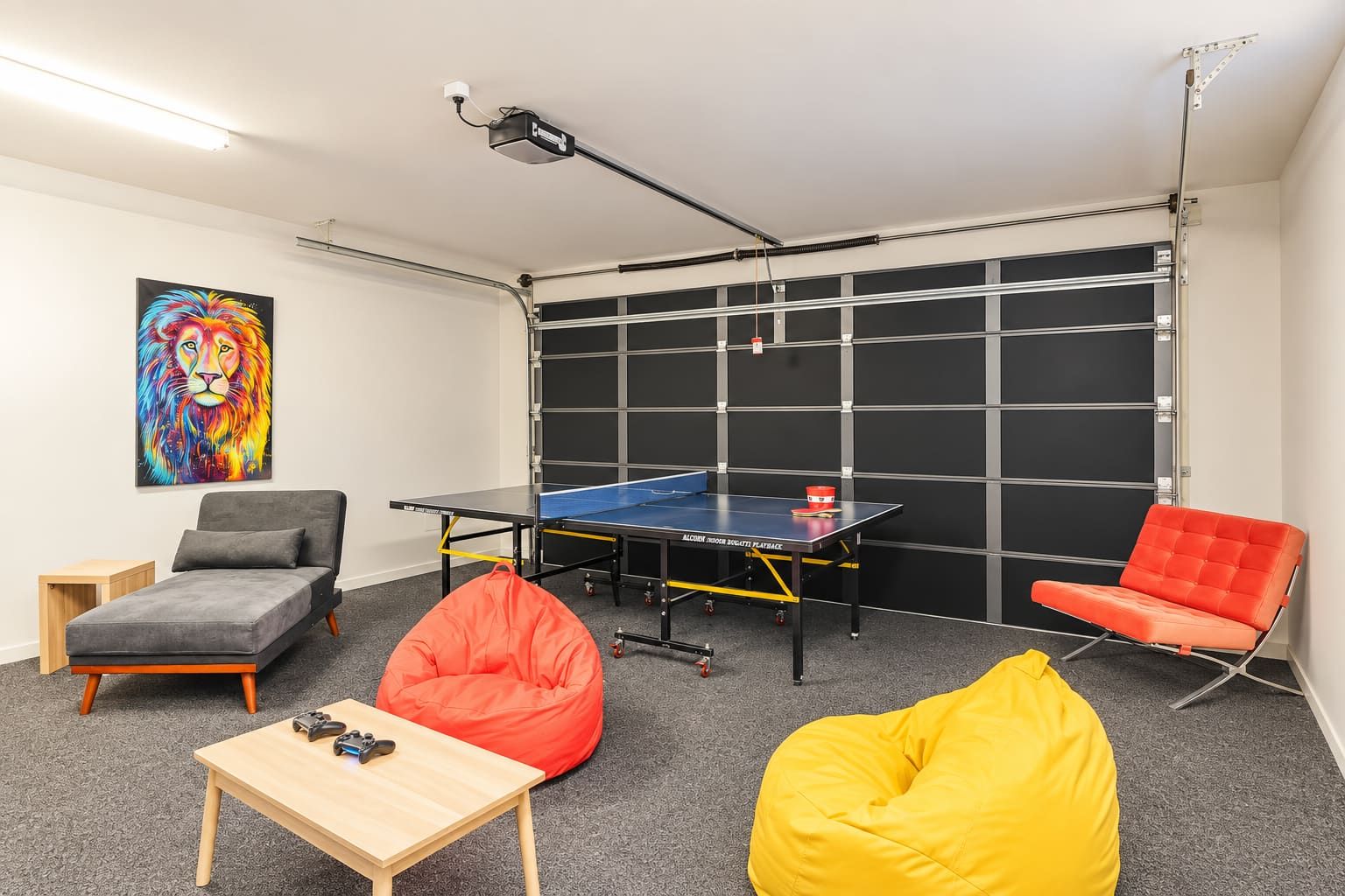 Ein Zimmer mit einem Pongtisch, zwei Sitzsäcken, einem Gaming-Sessel, einem kleinen Tisch mit einem Spielcontroller und einem farbenfrohen Kunstwerk an der Wand.