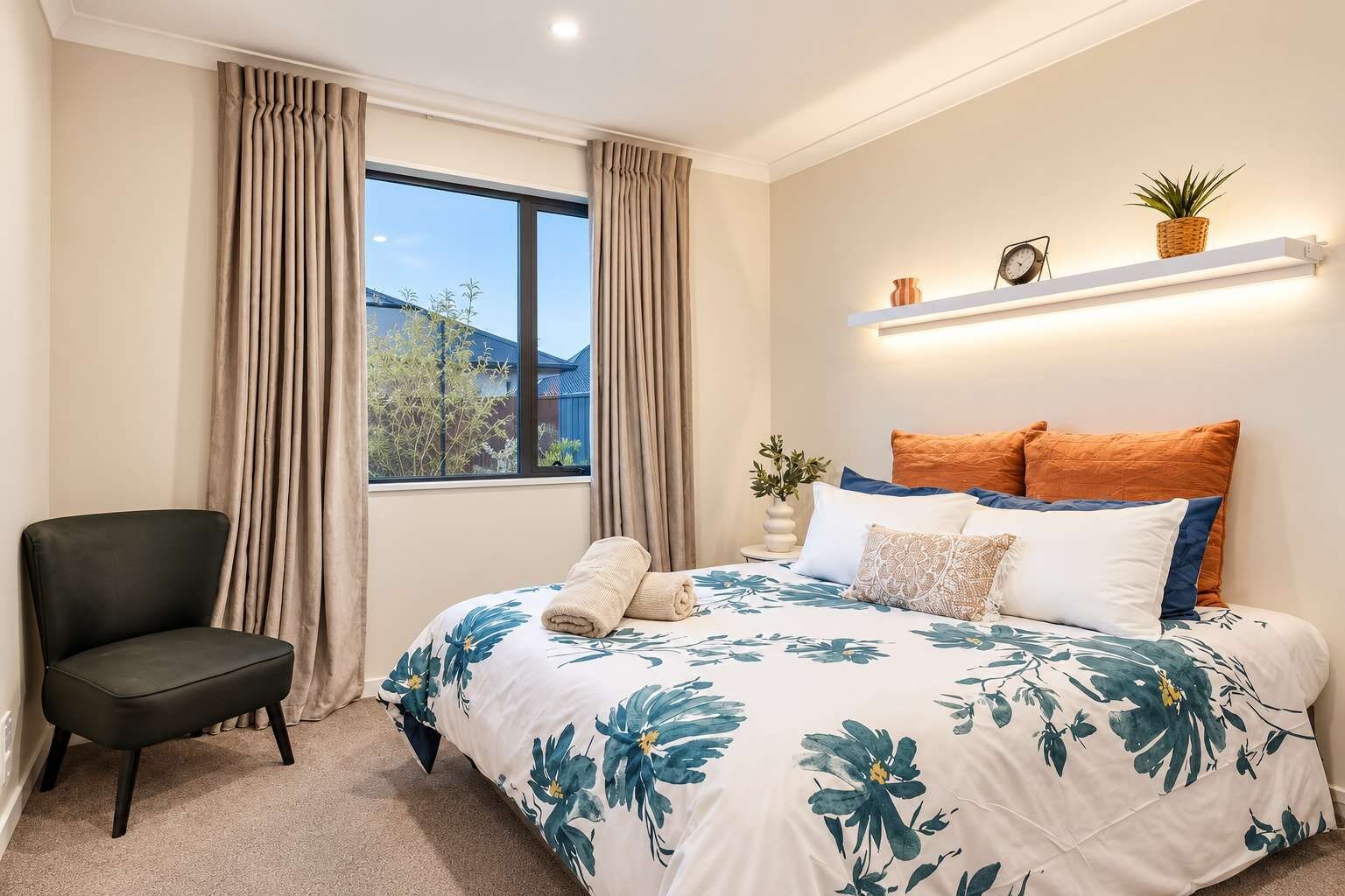 Een slaapkamer met een bed, een stoel, een raam met gordijnen, een nachtkastje met een plant en een plank met decoratieve spullen.
