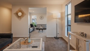 Un salon moderne avec un canapé, une table basse et un lampadaire. Un téléviseur est fixé au mur et une grande fenêtre laisse entrer la lumière naturelle.