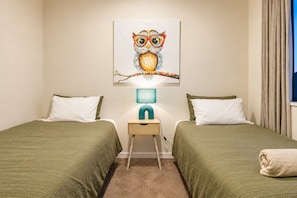 A room with two beds, a nightstand with a lamp, and a wall-mounted painting of an owl. - Wigram 3BR Home | Hot Tub + Games | Sleeps 8 (Christchurch)