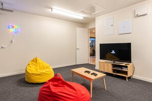 A room with a neon sign reading 'GAME ROOM,' a television, a wooden table with controllers, and bean bags. - Wigram 3BR Home | Hot Tub + Games | Sleeps 8 (Christchurch)