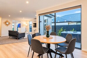 A modern dining area with a round table and six chairs, a television in the background, and a view of the backyard through large windows. - Wigram 3BR Home | Hot Tub + Games | Sleeps 8 (Christchurch)