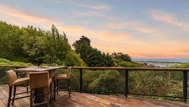 A wooden deck with a table and chairs, overlooking a coastal landscape with trees and a view of the sea. - Ocean Views | 3BR Home with Hot Tub & Parking (Christchurch)