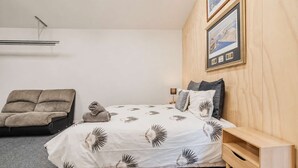 A bedroom with a bed, a sofa, a wooden nightstand, and a wall-mounted shelf. - Ocean Views | 3BR Home with Hot Tub & Parking (Christchurch)