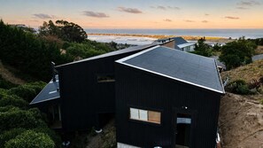 A modern black building with a sloped roof, surrounded by greenery and overlooking a coastal landscape. - Ocean Views | 3BR Home with Hot Tub & Parking (Christchurch)