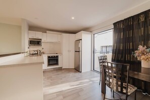 Een moderne keuken met witte kasten, een magnetron, een fornuis en een koelkast. Er is een eethoek met een tafel en stoelen, en een raam met gordijnen.