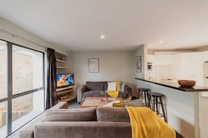 A modern living room with a sofa, a coffee table, and a TV. There is a kitchen area with a counter and stools. - Quaint Worcester Quarters - 3BR Delight (Christchurch)