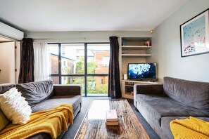 A modern living room with two grey sofas, a wooden coffee table, a TV on a wooden stand, and a large window with curtains. - Worcester 3BR | Garage | Central Christchurch (Christchurch)