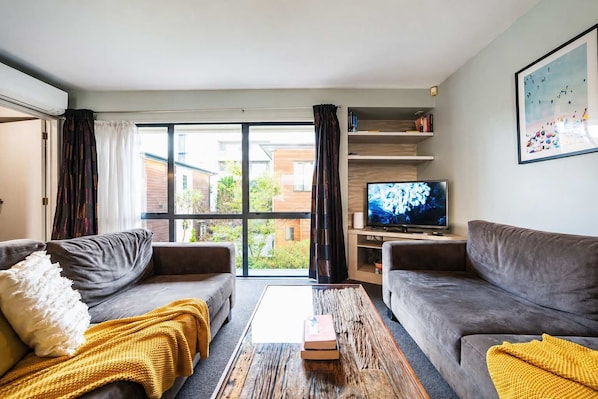 A modern living room with two grey sofas, a wooden coffee table, a TV on a wooden stand, and a large window with curtains.