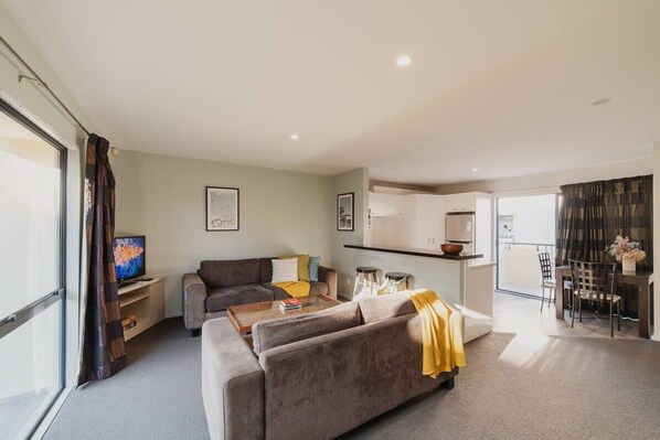 A modern living room with a sofa, coffee table, and a television. There is a kitchen area with cabinets and a dining table.
