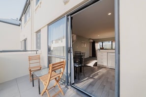 A modern apartment with a sliding glass door leading to a balcony with two wooden chairs and a table. Inside, there is a kitchen with white cabinets and a dining area with a black table and chairs. - Quaint Worcester Quarters - 3BR Delight (Christchurch)
