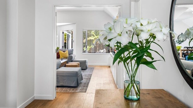 A hallway with a wooden floor, a glass vase with white flowers, and a mirror on the right. - KaleidoNook | 3BR + Parking + Private Yard (Christchurch)