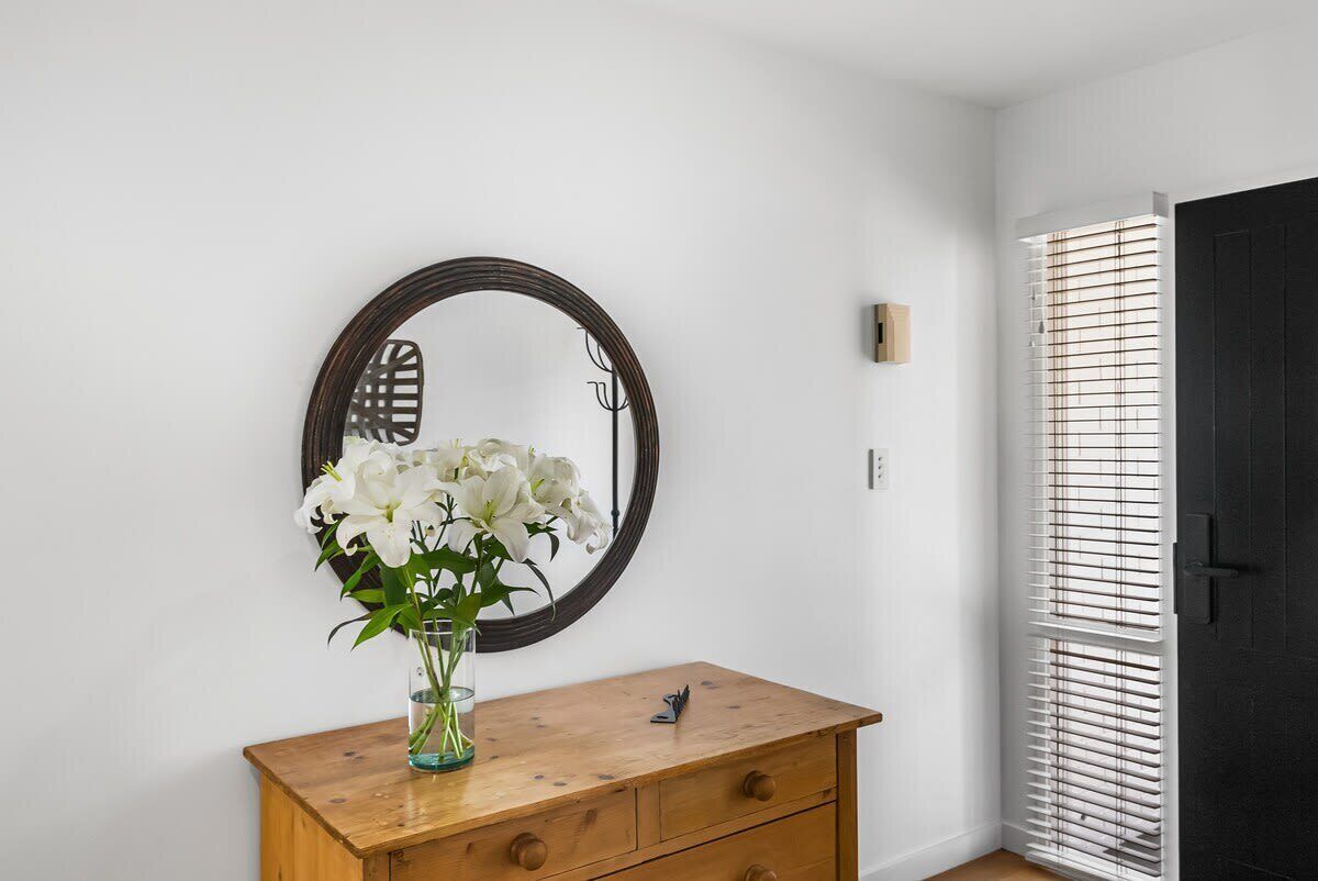 A wooden dresser with a vase of white flowers, a round mirror, and a black door with blinds.