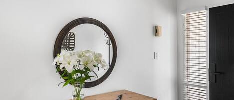 A wooden dresser with a vase of white flowers, a round mirror, and a black door with blinds.